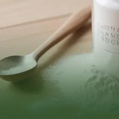 Cuchara de madera con polvo blanco junto a bote blanco sobre superficie clara, suplemento de colágeno